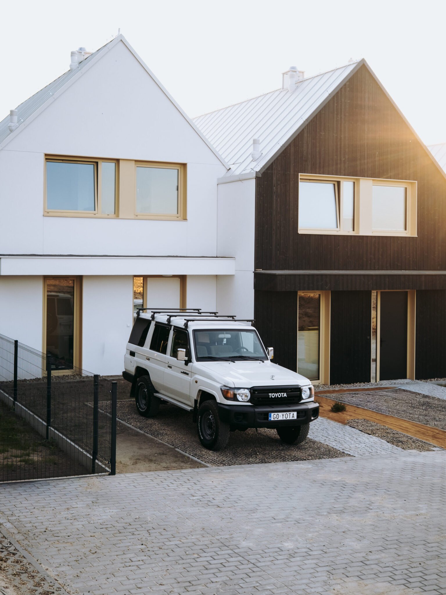 Land Cruiser 76 with Front Runner load bars parked on a gravel driveway in front of a modern house
